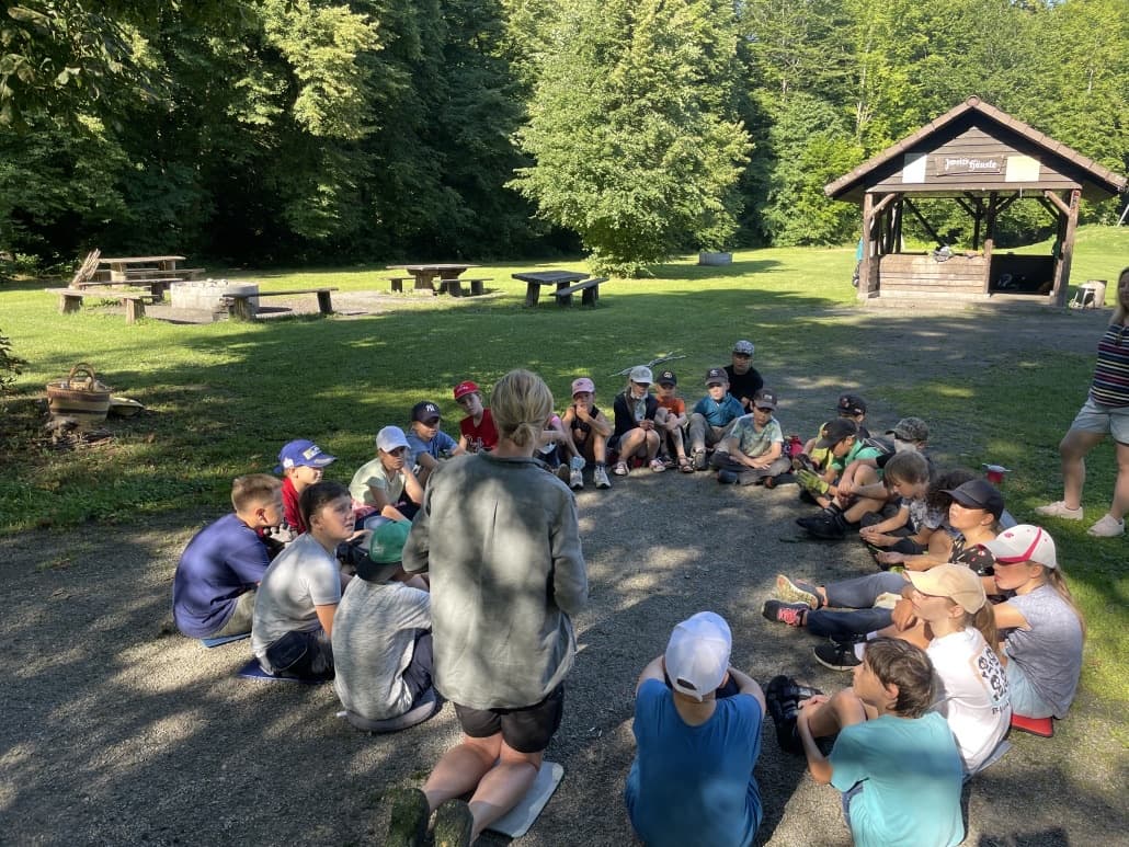 Kinder sitzen im Kreis vor einer Grillhütte im Wald